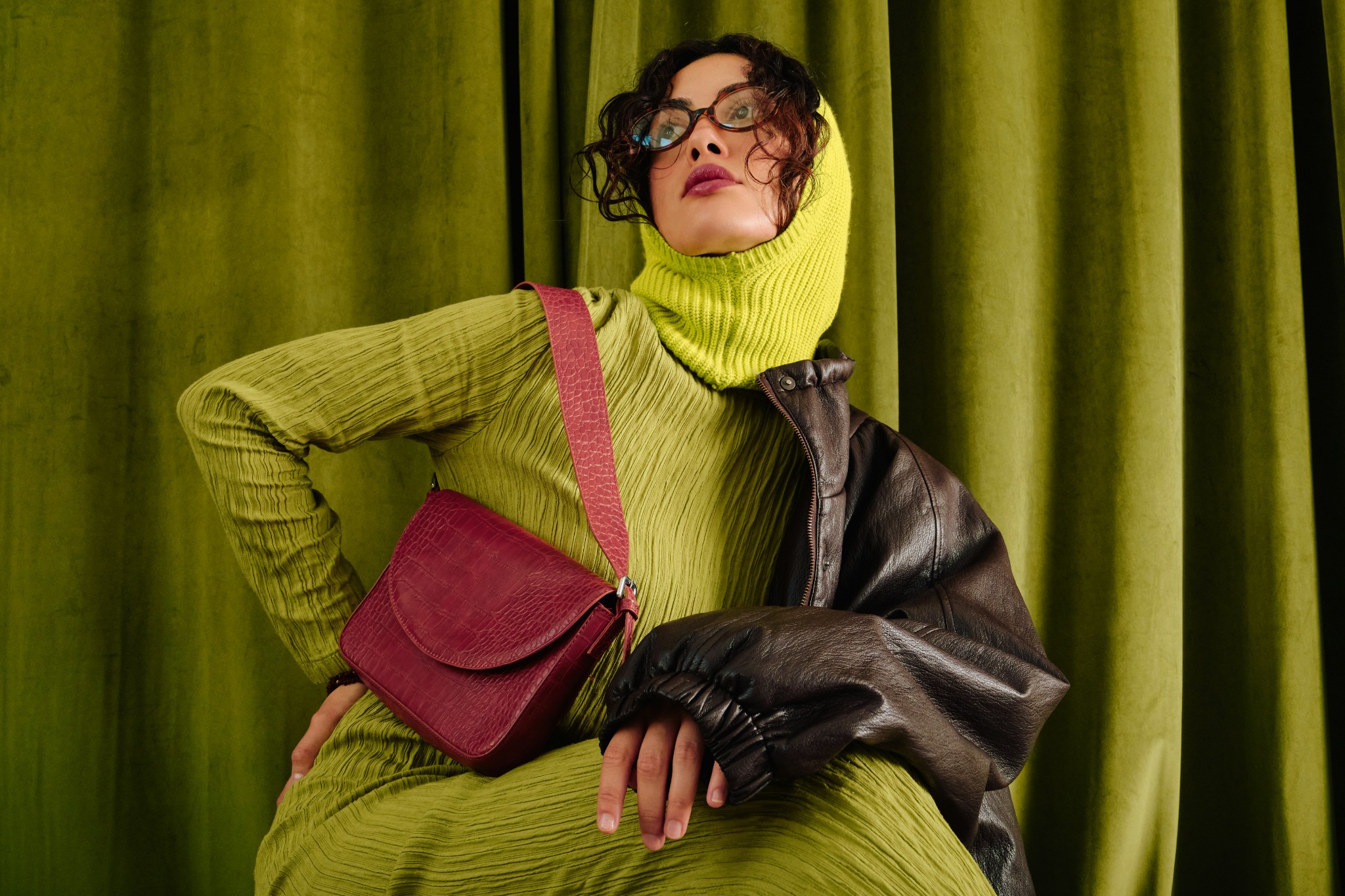 A fashion model wears a lime-green knit outfit with a burgundy shoulder bag against green curtains.