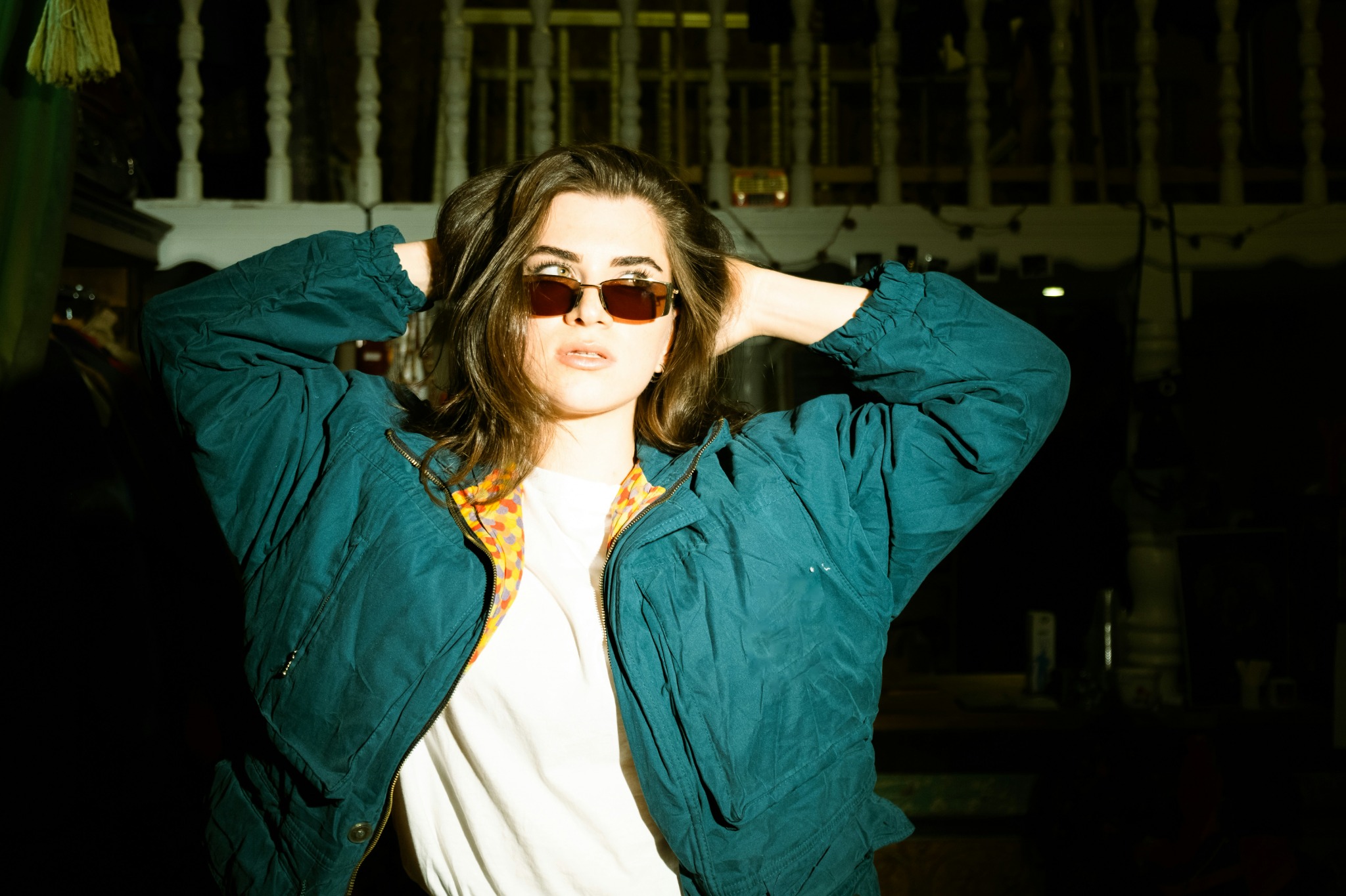 A model in a teal jacket and tinted sunglasses in a dark setting.